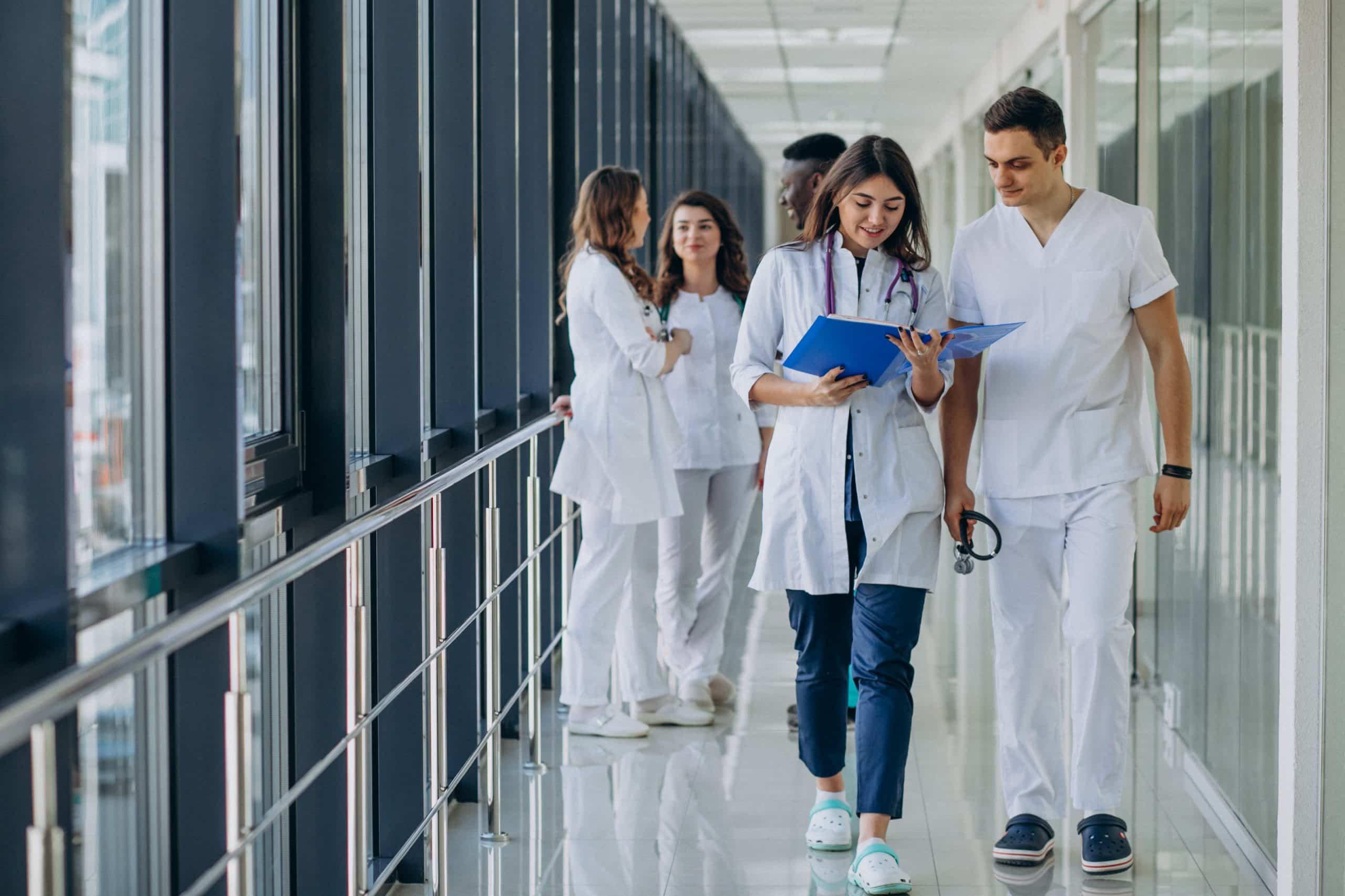 grupo de profissionais da enfermagem, de jaleco, caminhando em um corredor de hospital, sendo um homem e uma mulher no plano de frente da imagem e os demais ao fundo