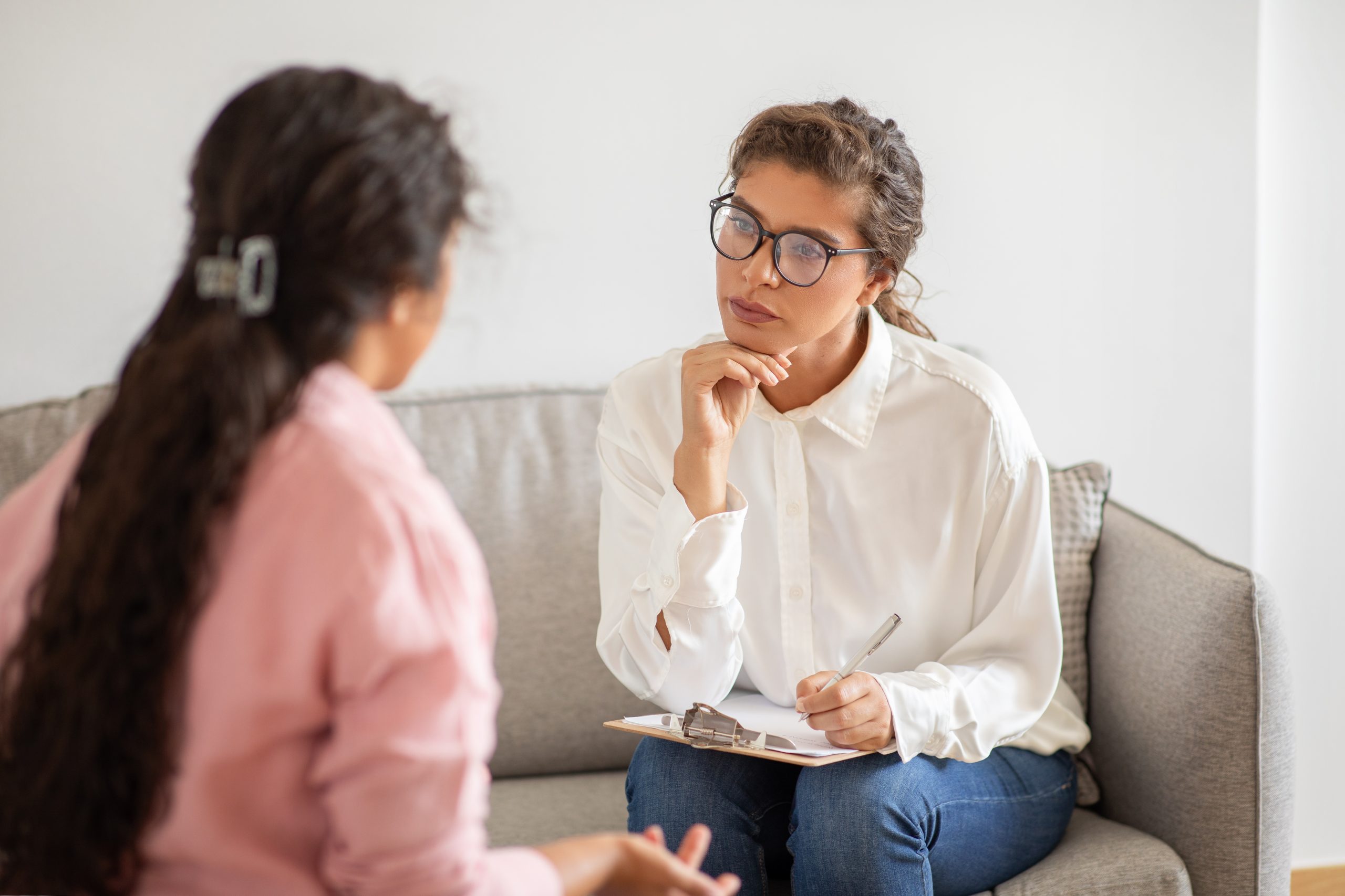duas mulheres sentadas frente a frente conversando. Uma delas está sentada no sofá com uma prancheta de anotações e a outra de frente para ela.