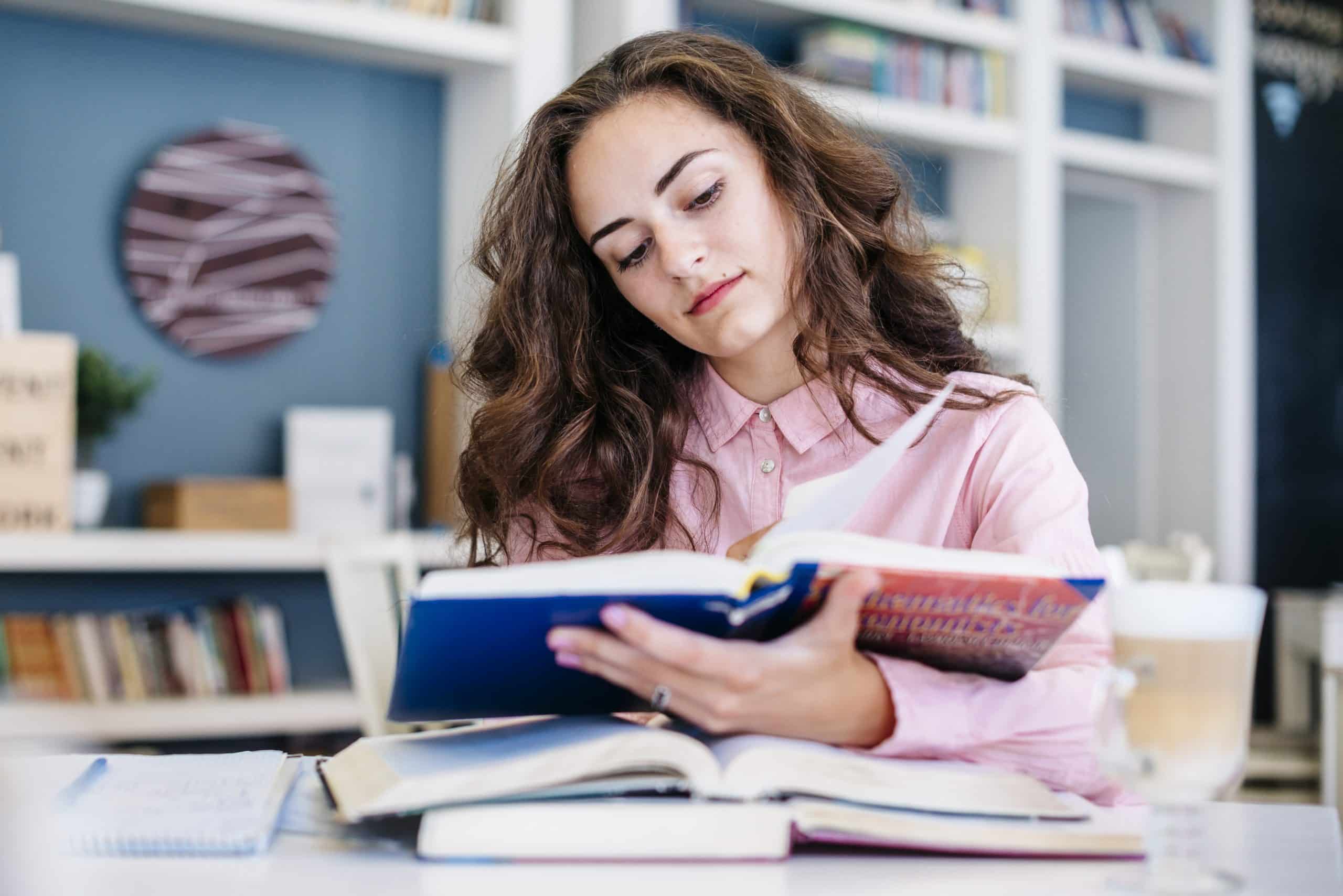 jovem adulta, sentada em uma biblioteca com três livros abertos, lendo um deles.