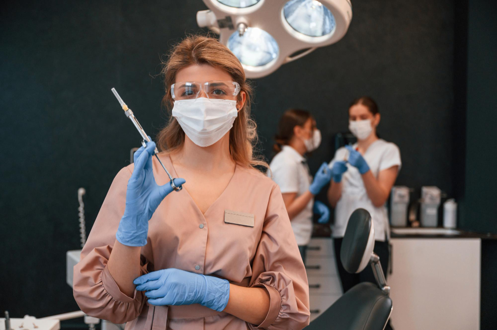 mulher com vestimenta de saúde e uma seringa na mão. Em segundo plano duas mulheres também com vestimentos em saúde, mexendo em produtos estéticos