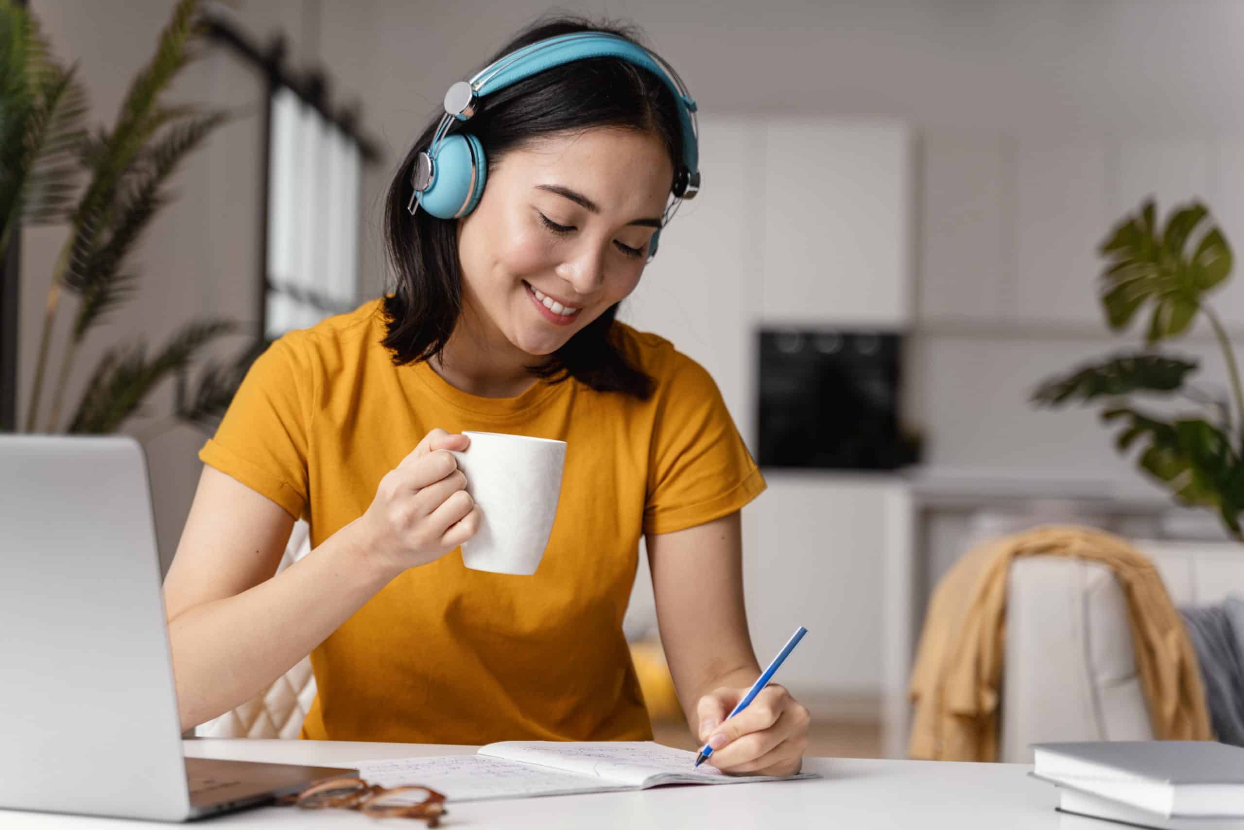 mulher sentada com fones nos ouvidos, segurando uma xícara de café e fazendo anotações em um caderno