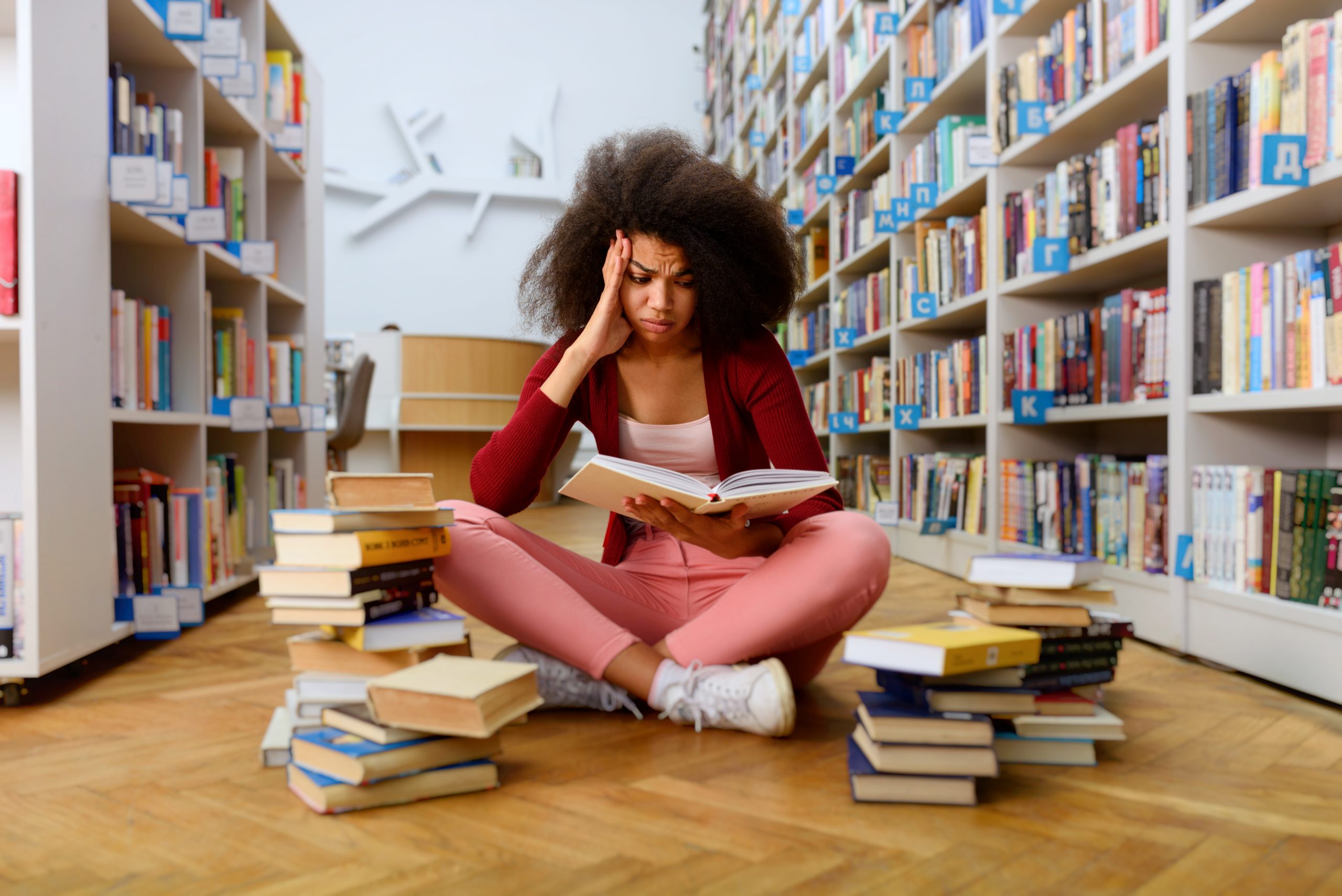 menina lendo, sentada no chão de uma biblioteca rodeada de livros junto dela. Sua expressão é de preocupada com conteúdo do livro