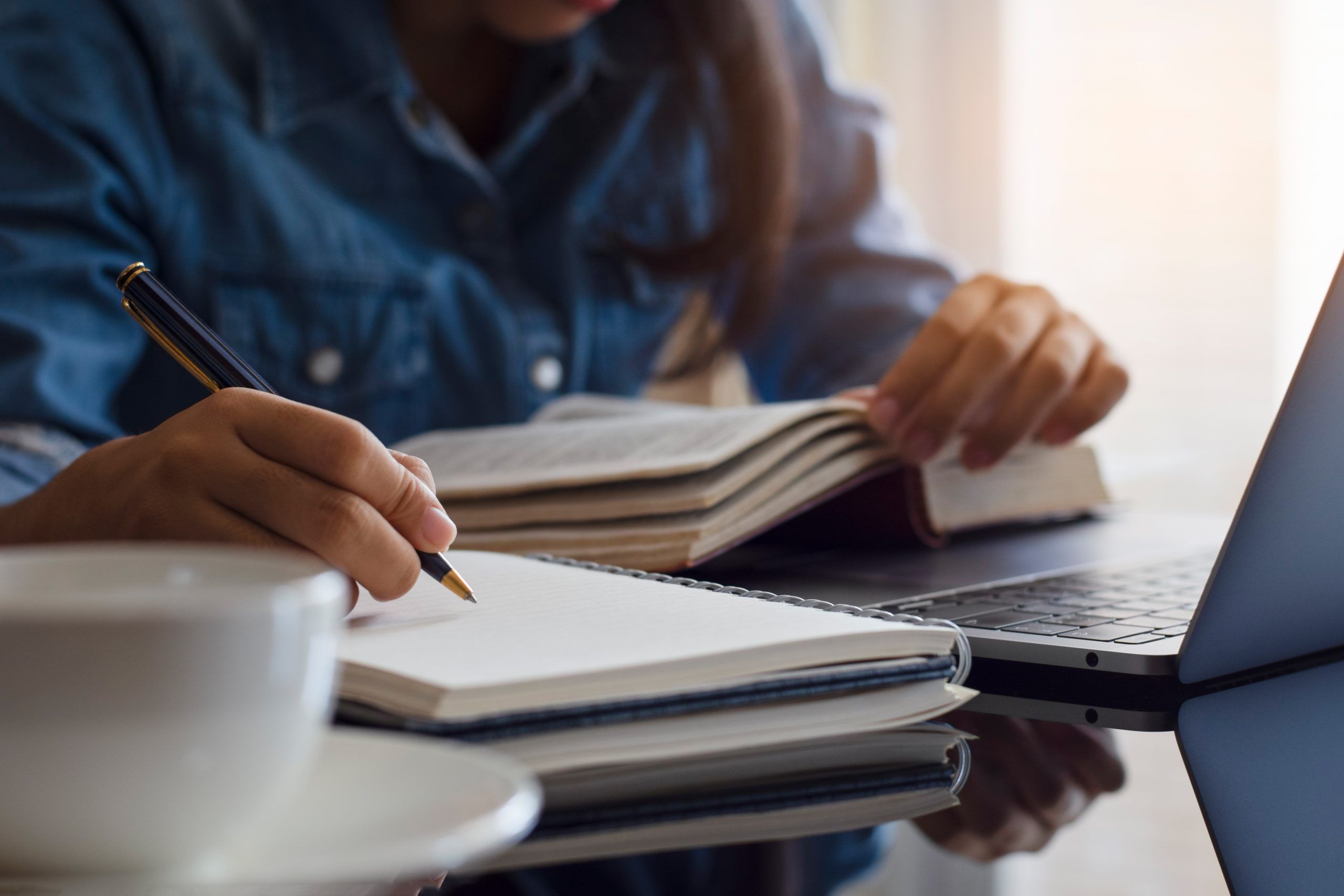 mulher estudando. Com uma mão sobre um livro e fazendo anotações com a outra em um caderno