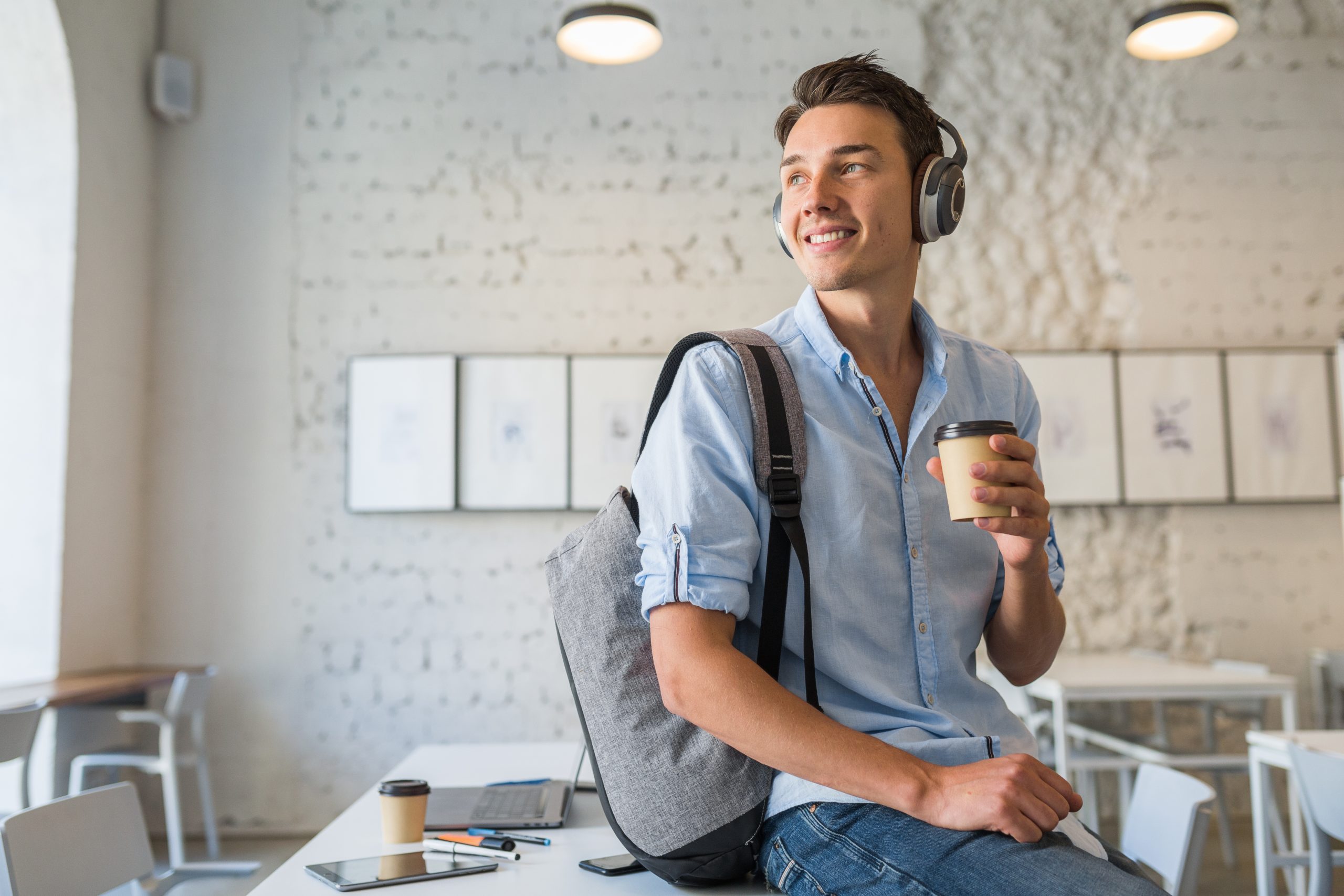 jovem adulto de fones de ouvido, mochila nas costas, segurando copo de café sentado em uma mesa.