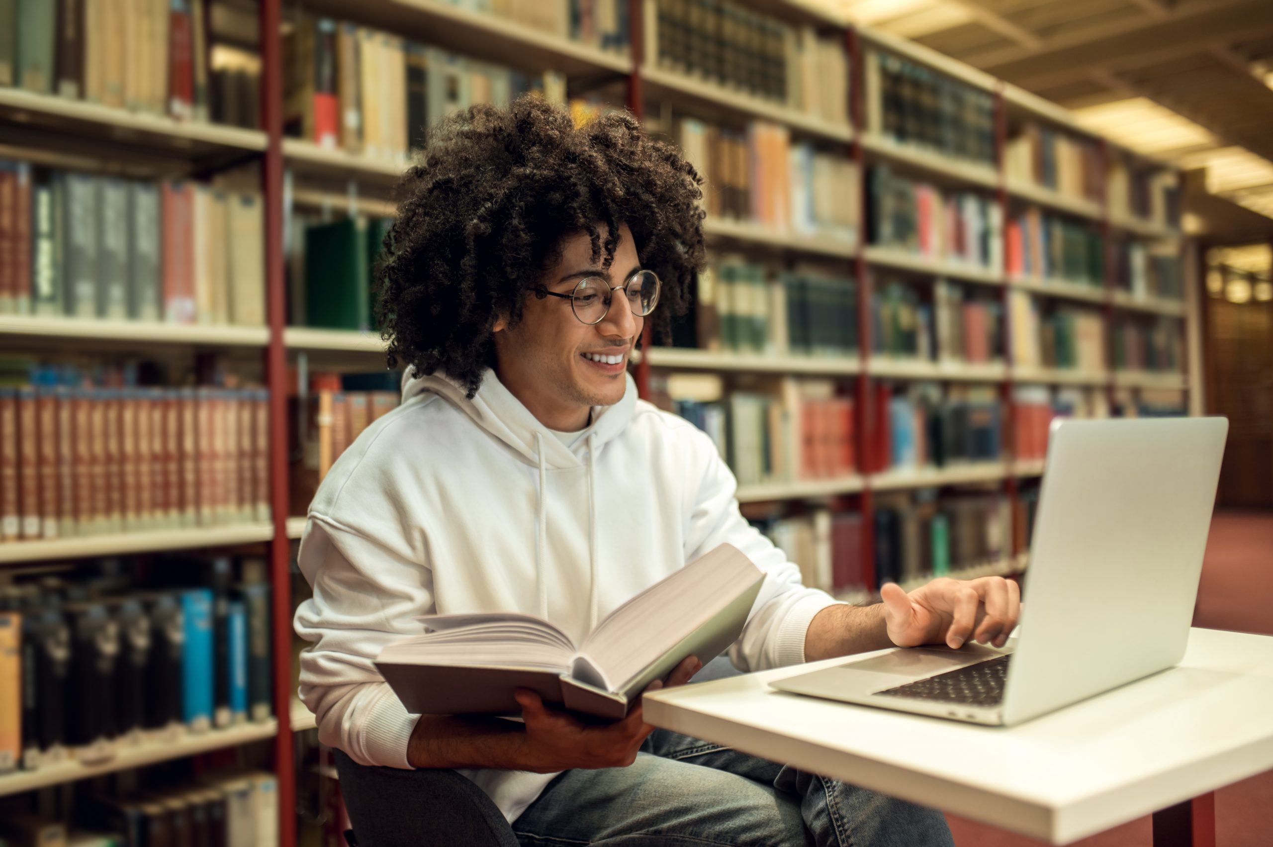 jovem adulto sentado em uma biblioteca com um notebook sobre a mesa e um grande livro em mãos.
