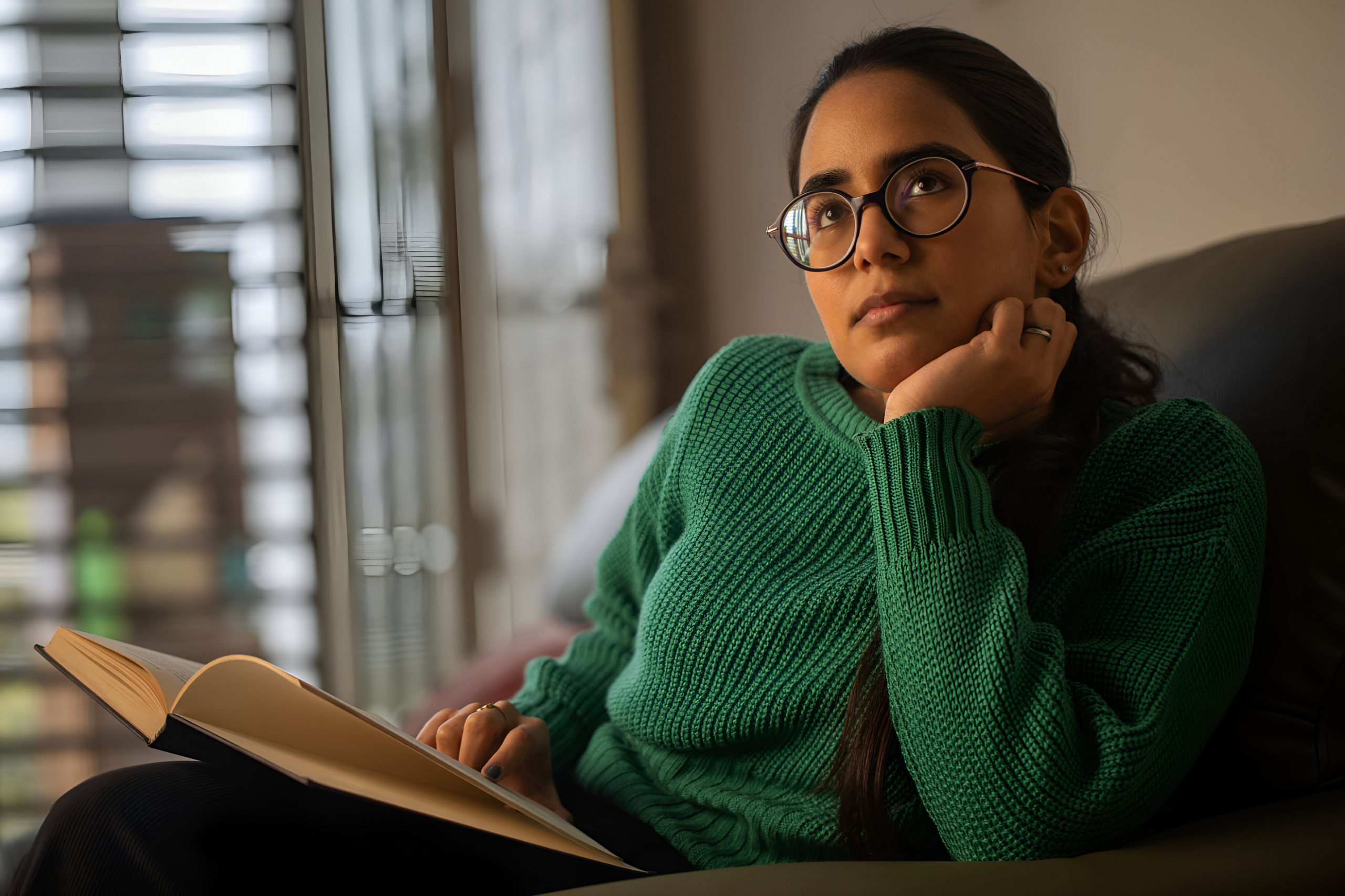 jovem adulta sentada no sofá com livro aberto em mãos, olhando para o horizonte pensando