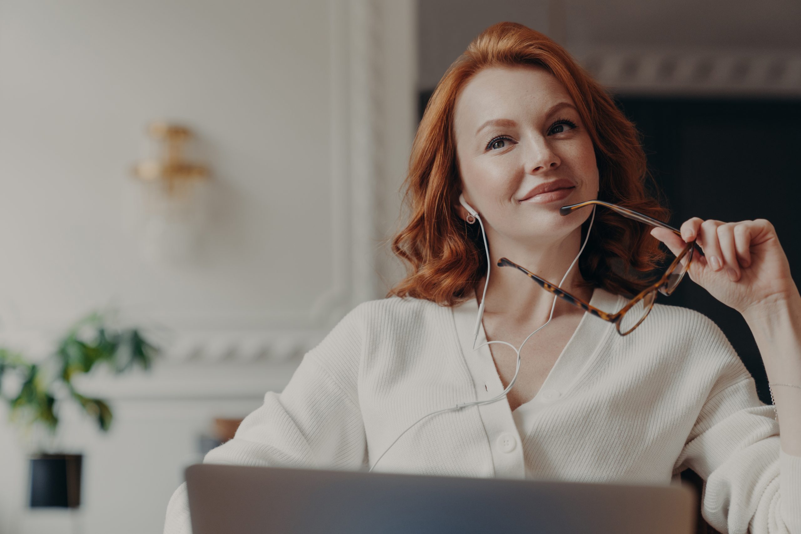 Mulher sentada em frente ao notebook. Ela está olhando para o horizonte, com fones nos ouvidos, apoiando os óculos na boca e com um leve sorriso no rosto. Sua expressão é de satisfação.