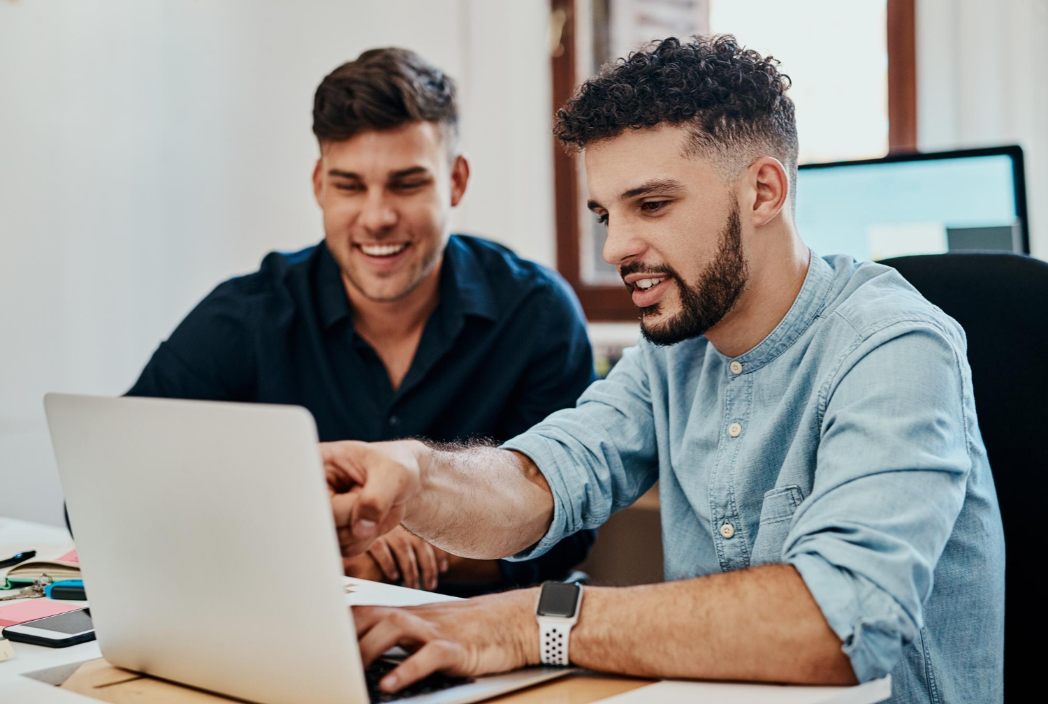 dois homens em escritório sentados em frente a um notebook, um deles está mostrando algo na tela para o outro, como se estivesse ensinando algo.