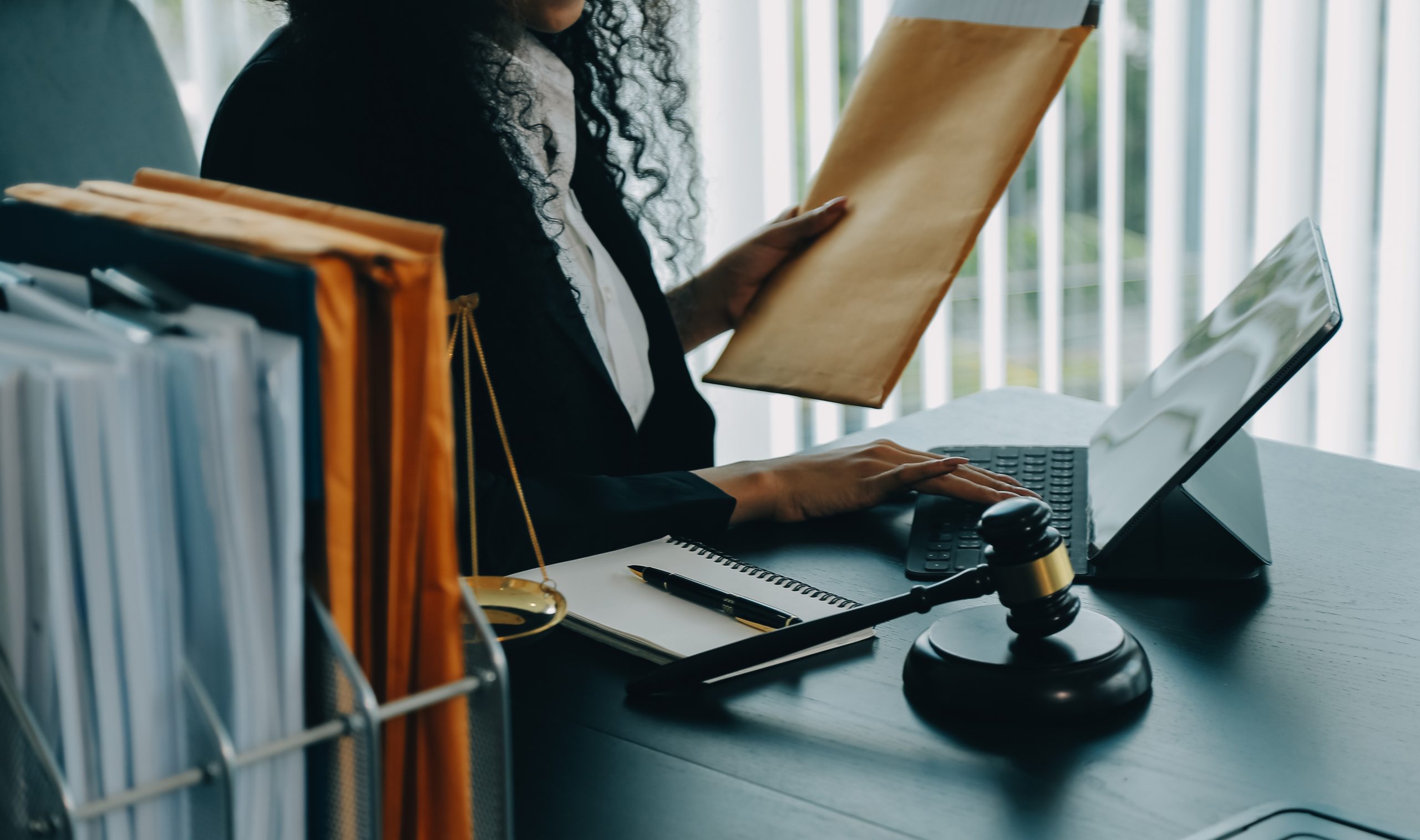 mulher sentada em um escritório de advocacia com um envelope em uma das mãos e mexendo no notebook, ao que parece fazendo uma consulta processual. Ao seu lado na mesa um gaveteiro com envelopes, um caderno com uma caneta em cima e um malhete.