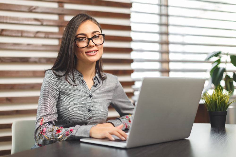Mulher sentada em ambiente profissional, com seu notebook na frente sobre a mesa, sorrindo para a câmera.