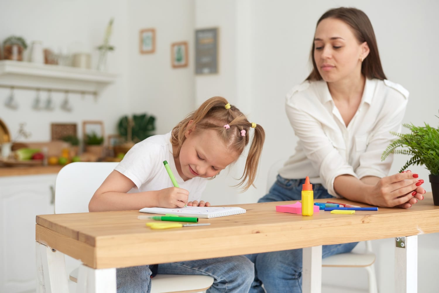 psicologa sentada ao lado de uma criança que está desenhando com canetinhas coloridas num caderno sobre uma mesa