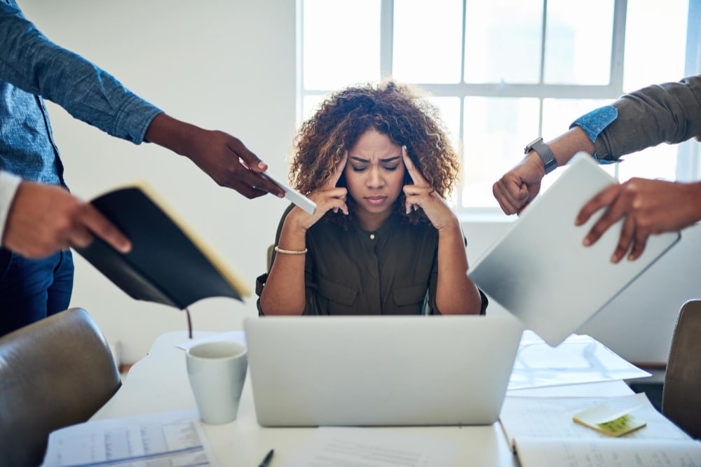 imagem de uma mulher sentada ao centro de uma mesa, com notebook em sua frente. Ao seu lado, aparecem diversas mãos de pessoas trazendo solicitações de trabalho, documentos, etc. Ela está com as mãos nas têmporas e de olhos fechados, com expressão de cansaço.