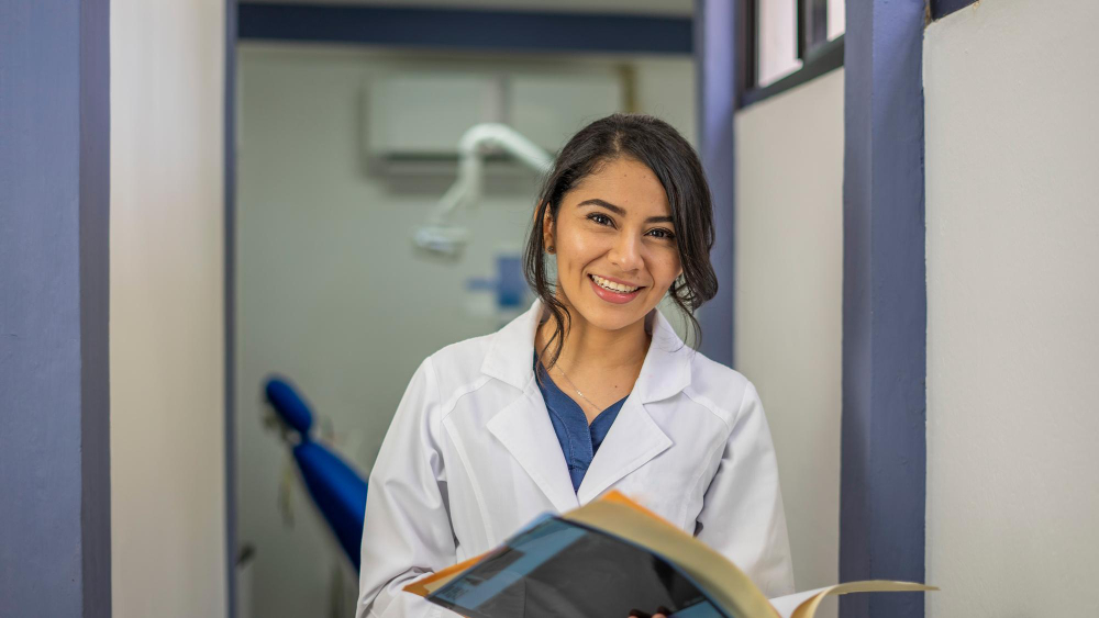 Mulher com jaleco de dentista em ambiente odontológico sorrindo para a câmera, ela está segurando um livro em mãos.