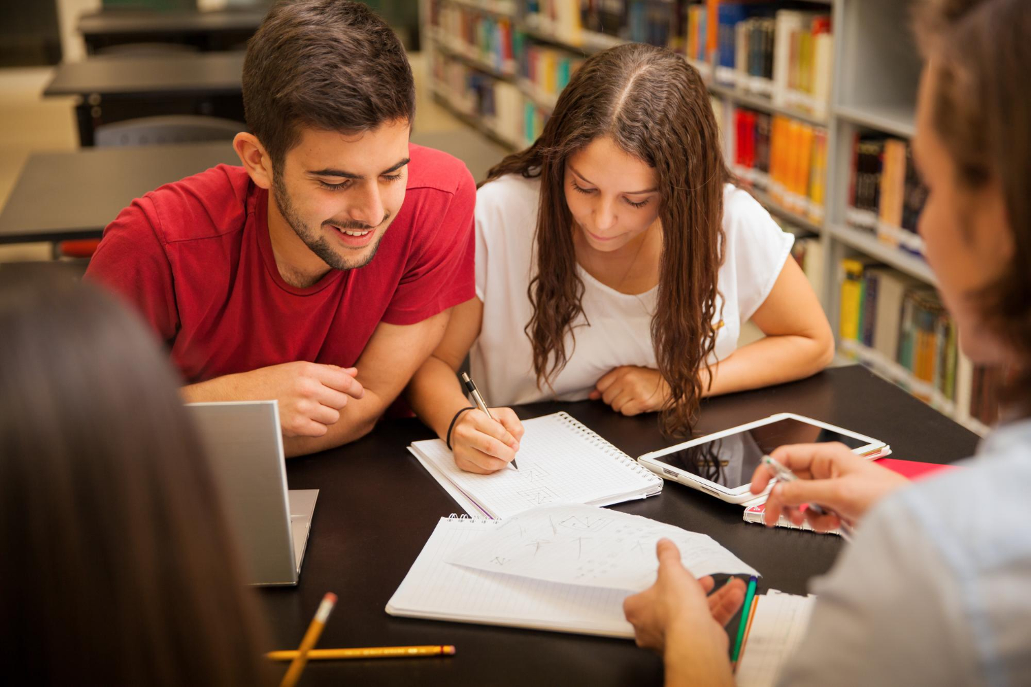 grupo de alunos estudando na biblioteca da faculdade. Sobre a mesa há um notebook, tablet e cadernos