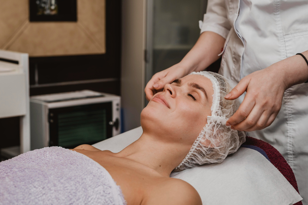 Mulher deitada em uma maca, enrolada em uma toalha, com touca estética enquanto outra profissional está atras dela para um procedimento