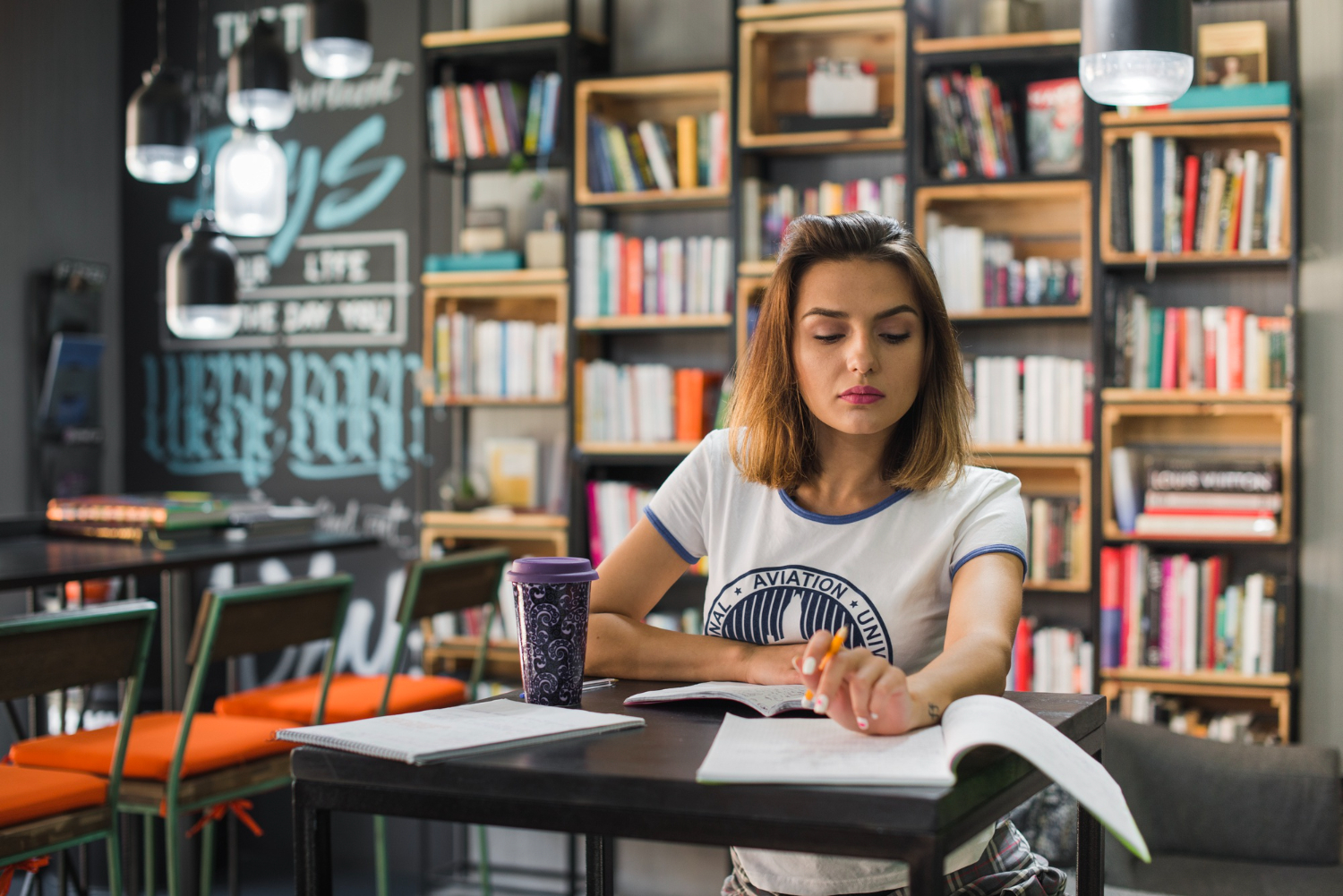 jovem adulta sentada na biblioteca estudando concentrada, com cadernos e livros abertos sobre a mesa