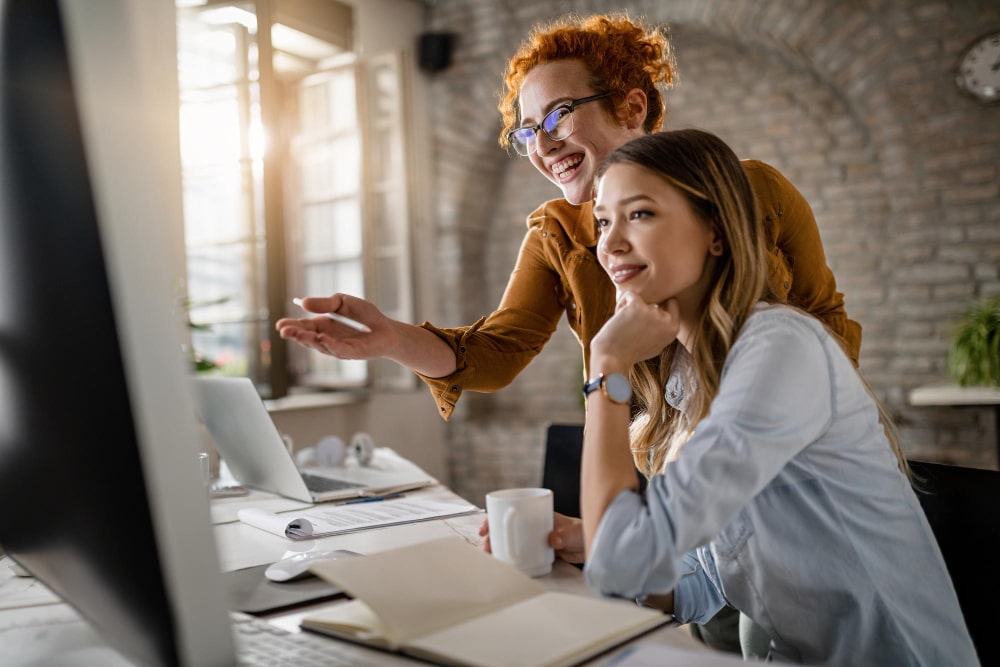 duas mulheres sorrindo em frente ao computador. Elas estão em ambiente de trabalho, com cadernos e anotações sobre a mesa