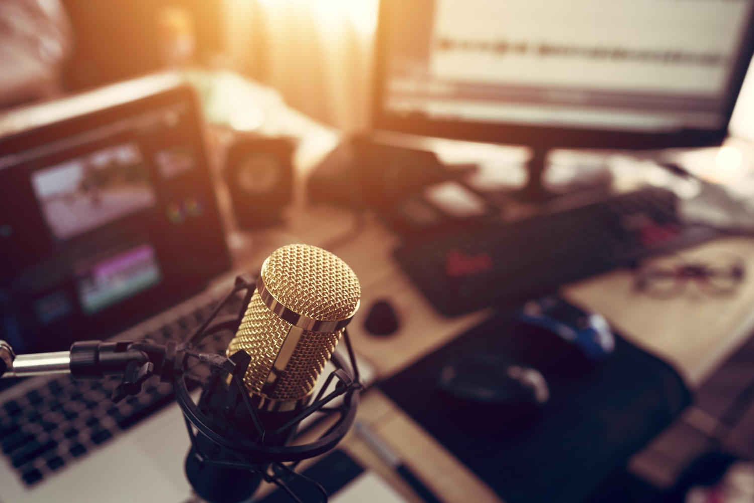 sobre uma mesa há dois computadores com sistemas de rádio e tv abertos, também um microfone. A imagem representa a produção ou apresentação de radiojornalismo