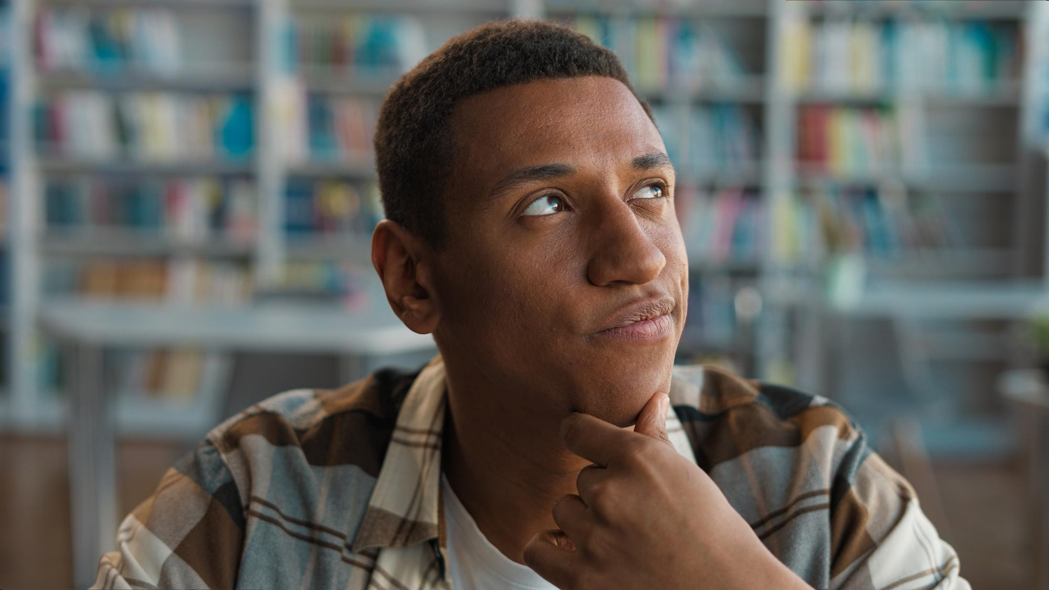 Jovem em uma biblioteca com a mão no queixo e semblante de dúvida, enquanto escolhe entre uma faculdade presencial ou EaD