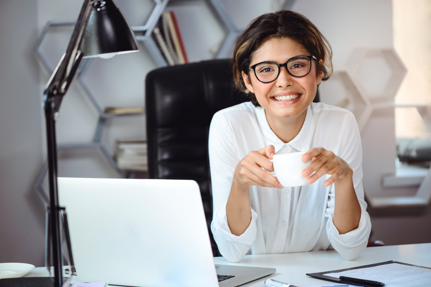 mulher sorrindo para a câmera. Ela está em ambiente de estudos, sentada em frente a um notebook e segurando uma xícara branca com as duas mãos.
