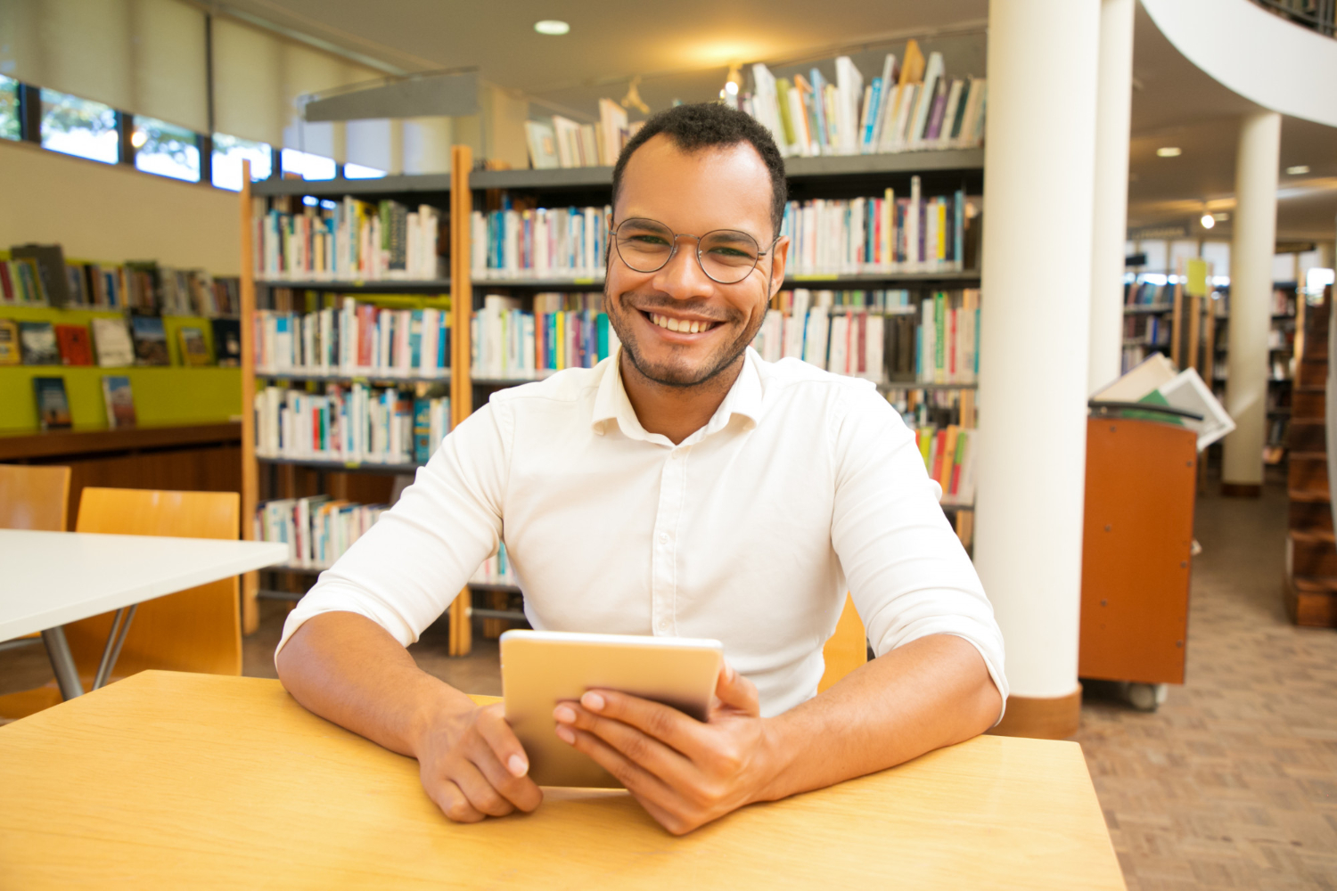 homem sentando sorrindo para a câmera. Ele está com camisa social, segurando um tablet sobre uma mesa de biblioteca. É possível identificar o cenário por conta da estante de livros ao fundo da imagem