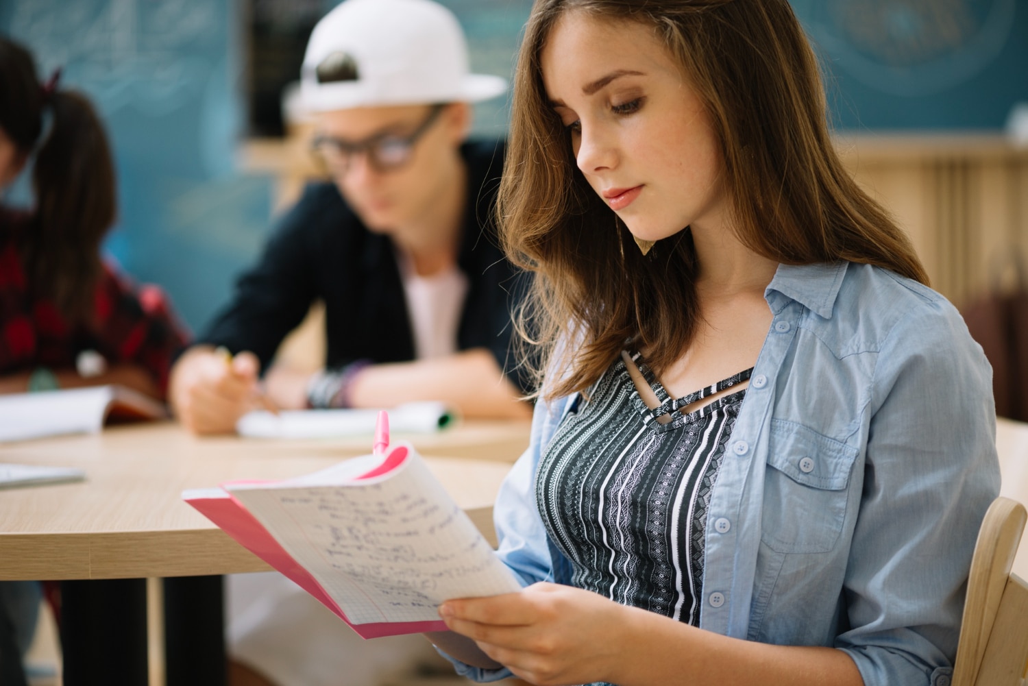 jovem adulta escrevendo em um caderno. Ela está em sala de aula, atrás há um menino também escrevendo em um caderno